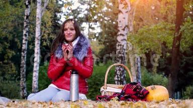 秋天公园里坐在野餐上喝热水壶里的热茶的年轻冷女人。坐在地毯上的女孩